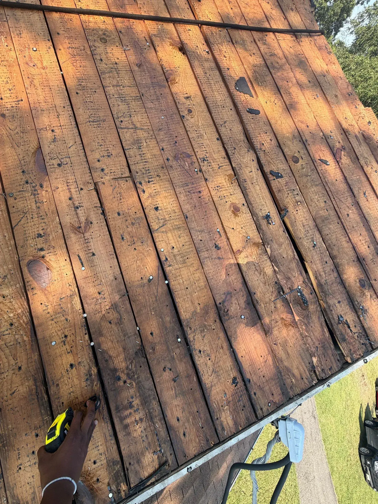 Wide sweep of bare plank decking studded with old nails, a hand holding a tape measure at the lower left